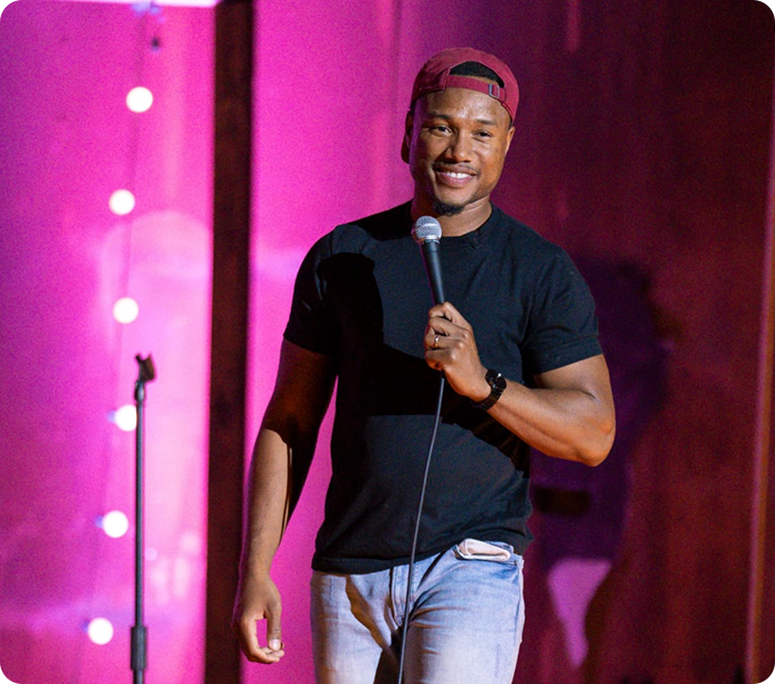 Person in hat on stage with microphone, pink background.